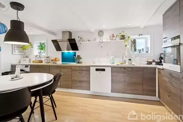 Et lyst køkken med træskabe, opvaskemaskine og emhætte; en bordplade med redskaber og planter står langs væggen. Rundt spisebord med sorte stole; blå og sorte pendellamper hænger fra loftet.