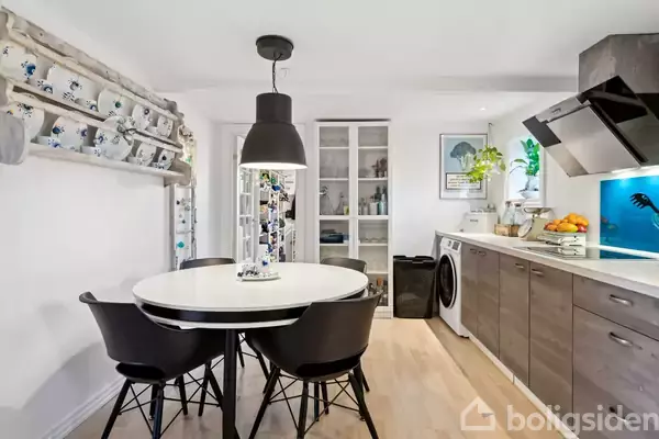 Rundt spisebord med sorte stole står i et lyst køkken. Bagved ses skab med glaslåger, og til højre er en vaskemaskine indbygget i køkkenbordet. Vaskemaskinen er omgivet af frugtskål.