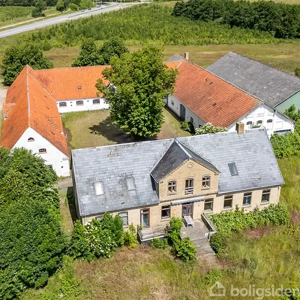 Et stort landhus med grå tagsten står omgivet af grønne marker. Til venstre er bygninger med røde tage, mens en grusvej fører gennem området. Træer omkranser stedet.