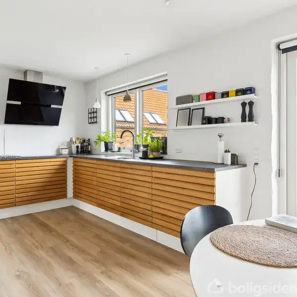 Et moderne køkken med træskabe langs væggene, en fladskærm monteret over arbejdsområdet, og en rund spisebord i forgrunden. Store vinduer giver udsigt til en have.