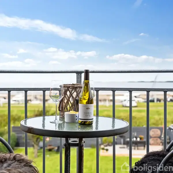 En flaske vin står på et rundt bord med et glas og en lanterne ved siden af. Bordet er placeret på en balkon med udsigt over en grøn mark under en klar blå himmel.