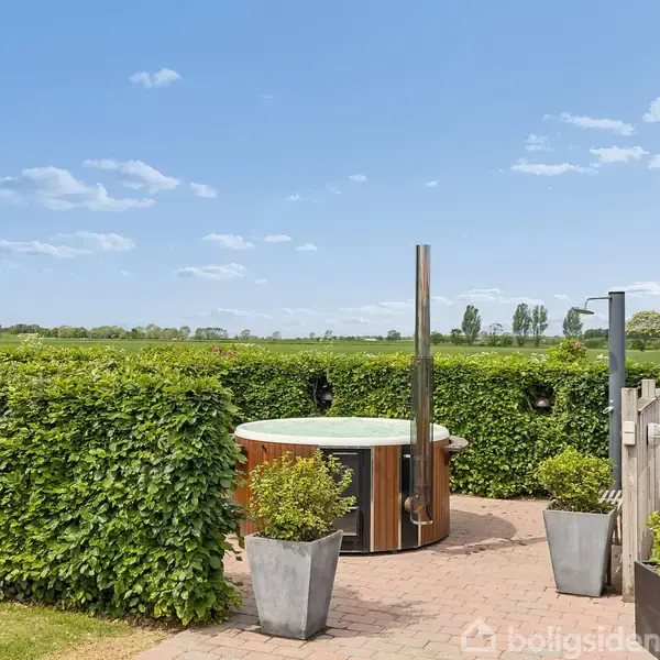 Jacuzzi står stille på en terrasse omgivet af hække og blomsterkrukker. I baggrunden ses en åben mark, omgivet af grønne træer under en blå himmel med spredte skyer.