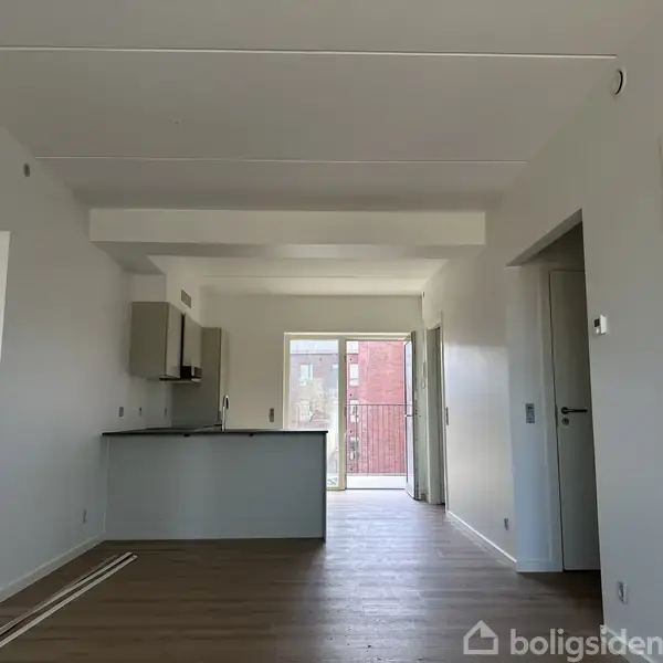 En moderne stue med trægulv fører til et åbent køkken med grå skabe. I baggrunden ses en stor altandør med udsigt til en rød murstensbygning.