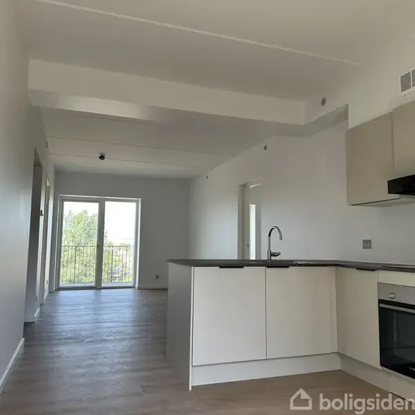 Et moderne køkken med hvide skabe og indbygget ovn står i et lyst rum med trælaminatgulv. Rummet har en stor glasdør, der fører ud til en balkon med udsigt.