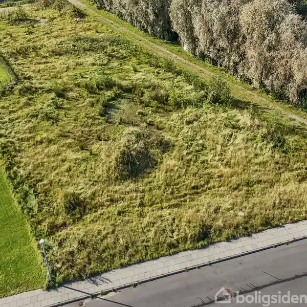 Tomt grund med højt græs ligger ubenyttet i et boligområde. Hække omgiver dele af grunden, og en asfalteret vej løber langs kanten.