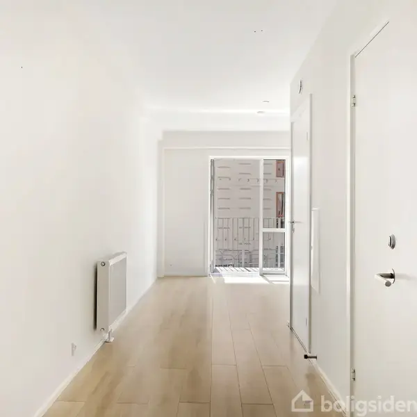 Et lyst og tomt rum med lyse trægulve strækker sig mod en glasdør, der fører til en balkon. En radiator er monteret på venstre væg, dør på højre.
