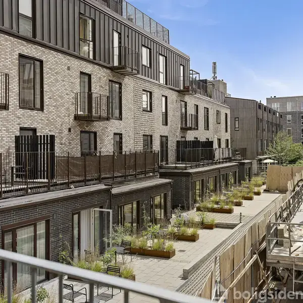 Flereetagers moderne boligejendom med murstensfacade, små altaner, og en række plantede krukker langs en gangsti. En byggekran og nabobygninger ses i baggrunden, under blå himmel.