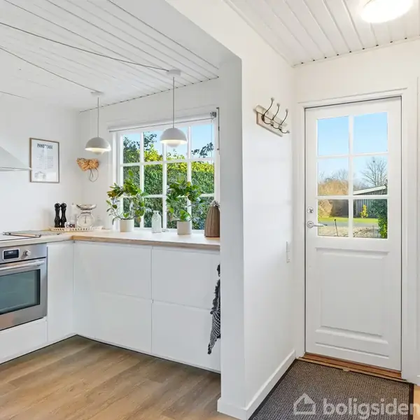 Et lyst køkken med hvide skabe og træbordplade indeholder en ovn. Over bordet er to hængende lamper. Der er et vindue og en glasdør med udsigt til en have.