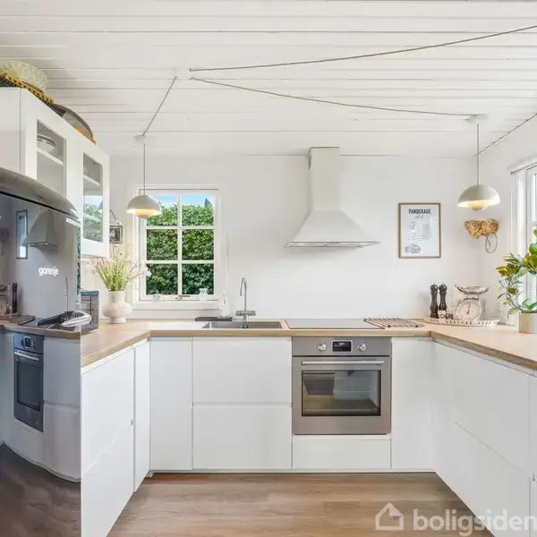 Køkken med hvide skabe og træbordplade, indeholder komfur og ovn. Køleskab til venstre, planter og dekorative elementer på bordpladen. Store vinduer giver naturligt lys og udsigt til grønne træer.