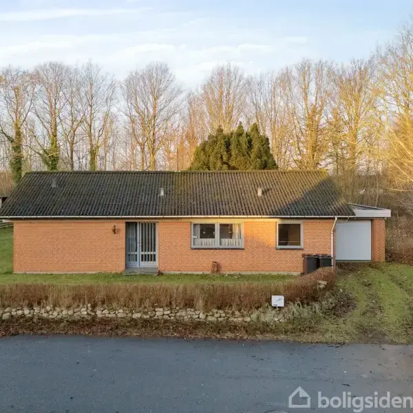 Et rødt murstenshus med sort tag står stille foran et græsareal og en vej. Baggrunden består af bartræer og løvtræer i vinterlandskab.