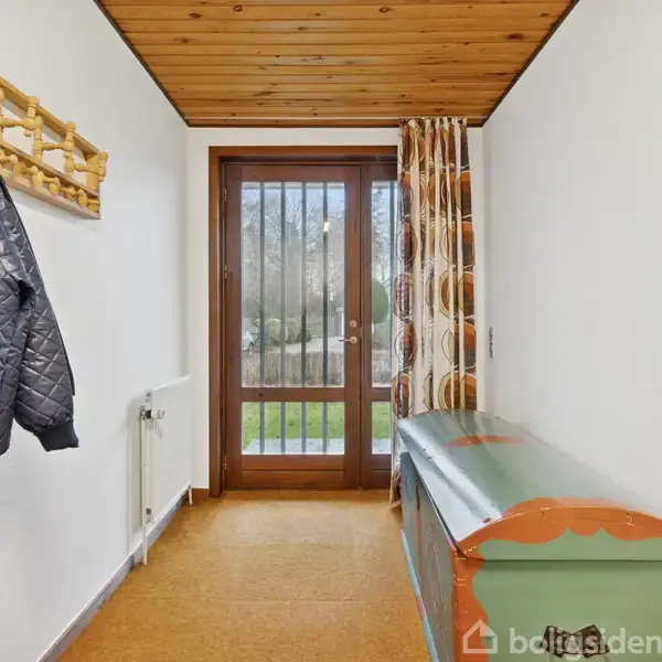 Jakke hænger på knagerække. Gangen har trægulv og brunt mønstret gardin ved glasdør, der fører ud til en have. En dekoreret trækiste står langs væggen.