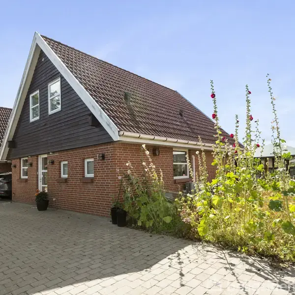Et rødt murstenshus med sort gavltag står ved siden af en asfalteret indkørsel. Blomster og grønne planter vokser op ad huset. Det er en solskinsdag.
