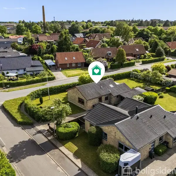Et parcelhus med mørkt tag står stille i et grønt boligområde omgivet af andre huse og veje. Et ikon markerer stedet på billedet.