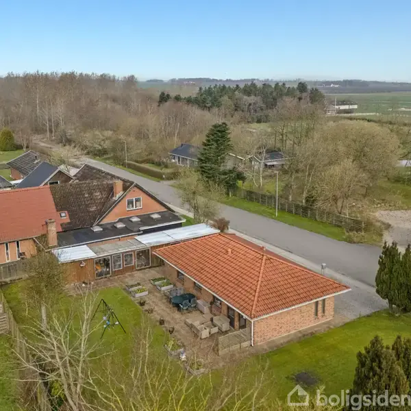 Et rødt tegltagshus med terrasse står stille i et landområde omgivet af træer, andre huse og en landevej.