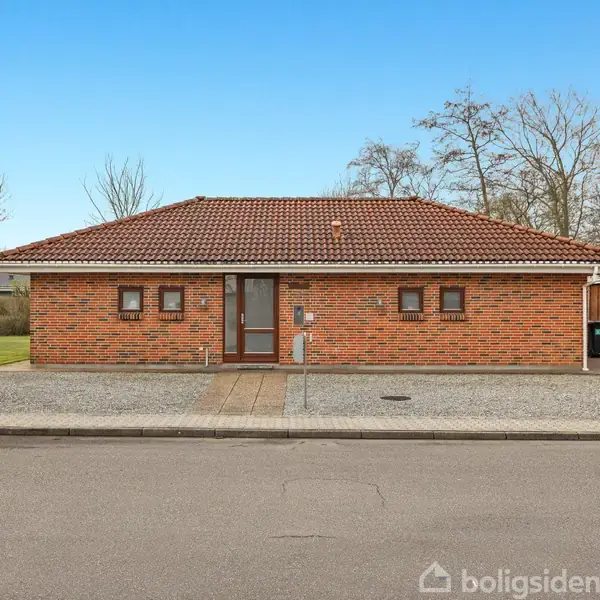 En enkeltstående murstensvilla med rødt tegltag står stille på en stille gade. Foran er der en grusbelagt indkørsel og græsplæne. Omkring ses træer og en carport.