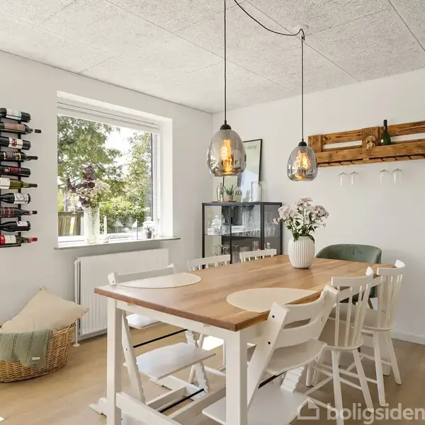 Et spisebord står i et lyst rum med hvide vægge og store vinduer. Bordet er omgivet af hvide stole, og en vase med blomster står på bordet. Væghængte vinreoler og en kurv ses i rummet.
