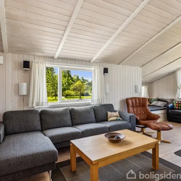 Gråt sofahjørne står foran stort vindue med udsigt til grøn have; ved siden står brunt lænestolssæt og sofabord i lyst træ; lyse trævægge og loft omgiver rummet.