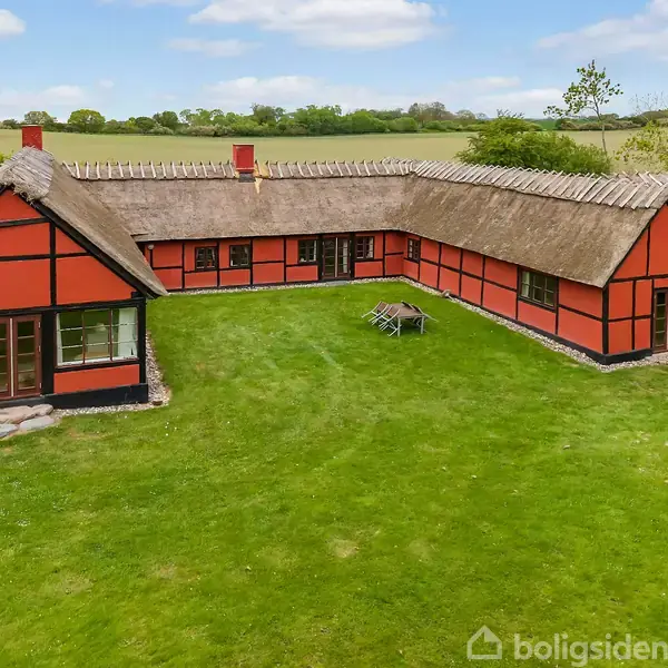 To røde, stråtækte bygninger ligger i grønne omgivelser ved siden af en vej. Omkring dem er en tæt skov med udsigt over en blå bugt i baggrunden.