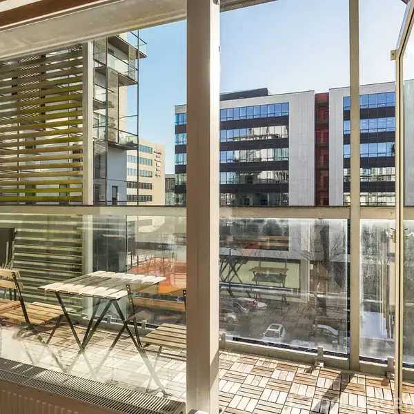 En altan med bord og stole set indefra, åbne glasdøre, i et moderne byområde omgivet af højhuse og lejlighedsbygninger.