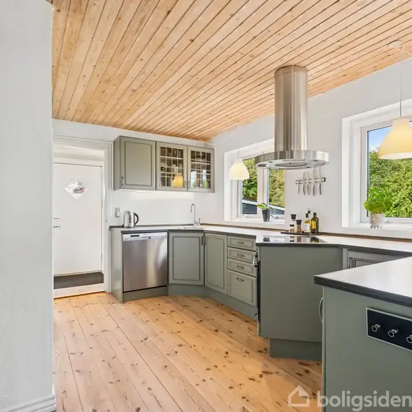 Et moderne køkken med grå køkkenskabe og en køkkenø under en lys træloft. Vinduerne slipper dagslys ind, og der er hængelamper over bordpladen. Udsigt til grønne træer udenfor.