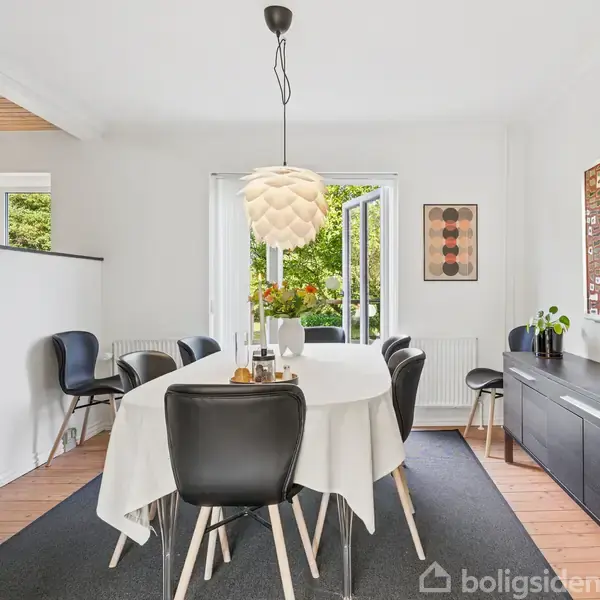 Spisebord med hvid dug omgivet af sorte stole står i et lyst rum. En moderne lampe hænger over bordet, og gennem et åbent vindue ses grønne træer udenfor.