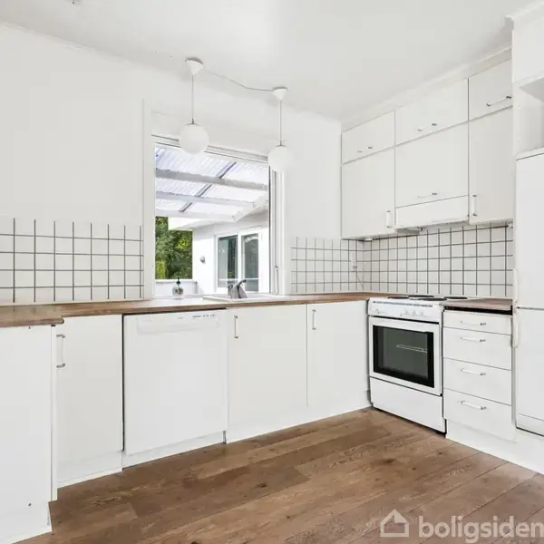 Køkken med hvide skabe og skuffer. Køleskab, ovn og opvaskemaskine står langs væggen. Der er lyse lamper over en træbordplade. Brune trægulve og et vindue med udsigt til grønne omgivelser.