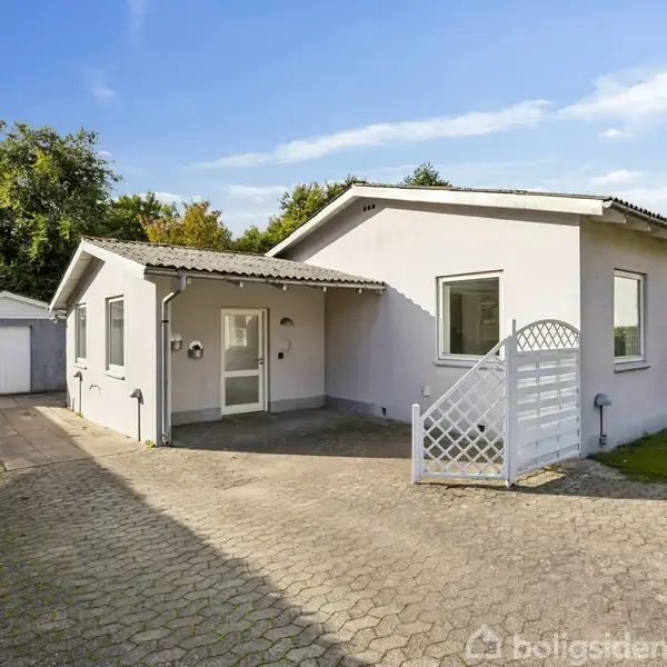 En hvid bolig med fladt tag står i en have med en indkørsel og garage til venstre. Skyfri blå himmel og grønne træer omkranser huset.