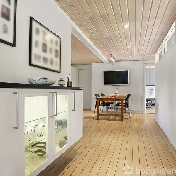 Skab med glaslåger viser porcelæn, mens glas og dekorationer står ovenpå. I baggrunden et spisebord med fire stole, hængende fladskærm på væg, lyst træloft og gulv. Ingen tekst.