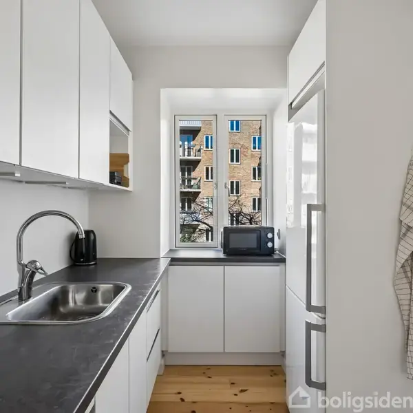 En hvidt køkken med sort bordplade har en vask og sort elkedel. En mikrobølgeovn står ved vinduet. Et viskestykke hænger ved et skab. Udenfor ses en bygning.