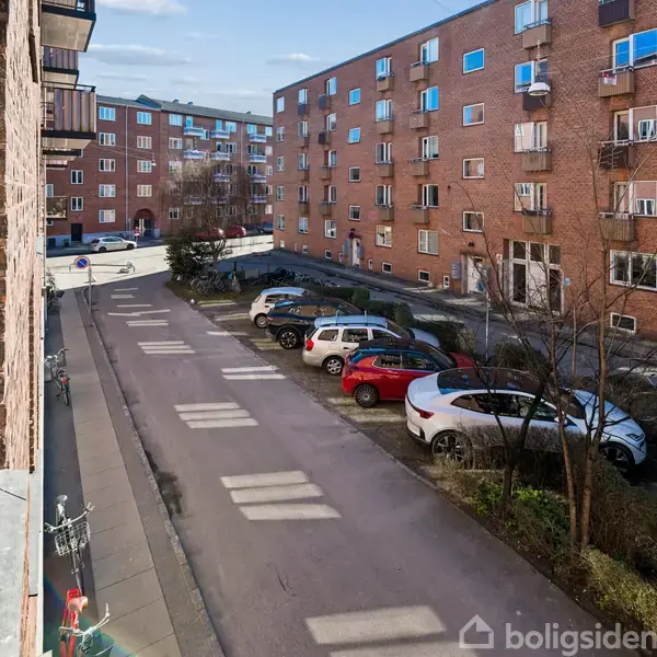 Et parkeringsområde med biler holder stille mellem flere murstensbygninger i et bykvarter. Fortove flankerer vejen. Skyer pryder himlen i baggrunden.