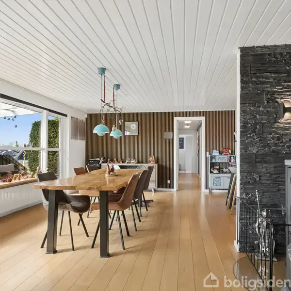Brændeovn i stue opvarmer hyggeligt rum. Spisebord med stole ved store vinduer. Moderne lysekrone hænger over bordet. Trægulve og -loft dominerer interiøret. Udendørs terrasse synlig gennem vindue.