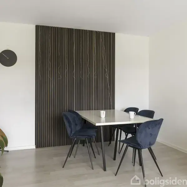 Et moderne spisebord med fire blå stole står i et lyst rum. I baggrunden er der en væg med dekorative trælister. Ved siden af står en grøn plante og en guitar.