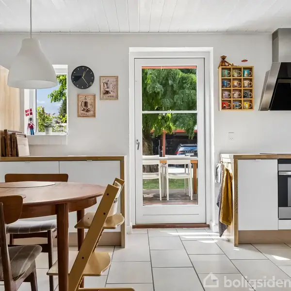 Et køkken med et rundt spisebord og stole, køleskab til venstre. På væggen ses ure og billeder. En glasdør fører til en terrasse med udsigt til træer.