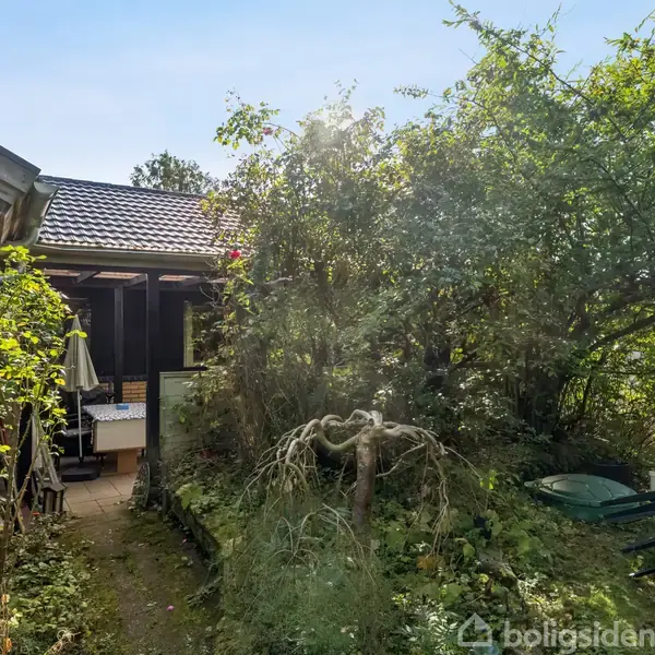 Haven med grønne buske og høje træer omgiver en terrasse med plads til siddepladser og en parasol. I baggrunden ses en del af et hus' murstensvæg.