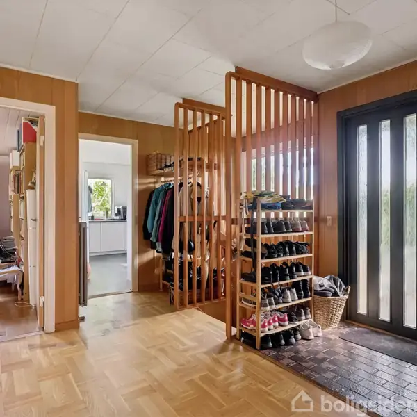 Entré med træhyldearrangement fyldt med sko, jakker hænger ved siden af. Store glasdøre slipper lys ind. Baggrund har synlige værelser med bøger og et vindue.