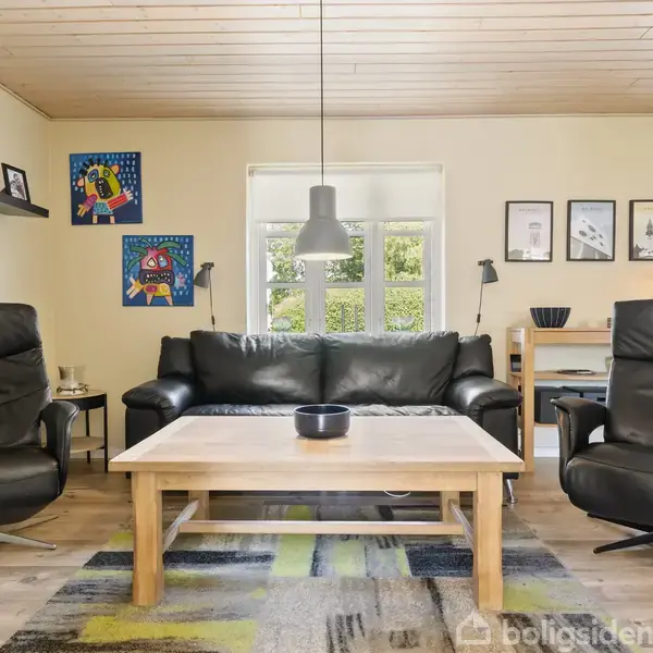 Sort lædersofa og to lænestole står omkring et træbord i en lys dagligstue. Malerier og billeder hænger på væggene; vinduerne giver udsigt til grønne træer.