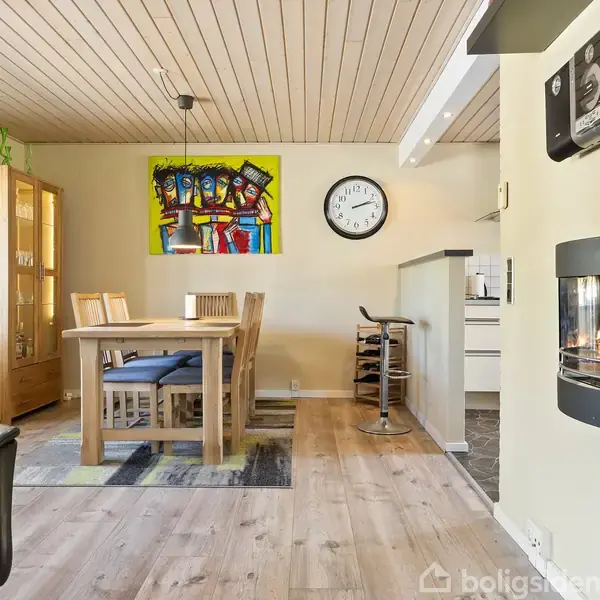 Et spisebord med seks stole står i et lyst rum med træloft. En farverig maleri og en ur hænger på væggen. En pejs er tændt.