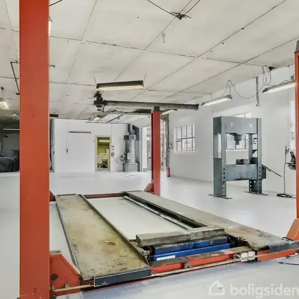 En bilværkstedslift står inaktiv i en hvidmalet garage med højt til loftet. Der er lysarmaturer i loftet og en brandslukker placeret ved siden af liften.