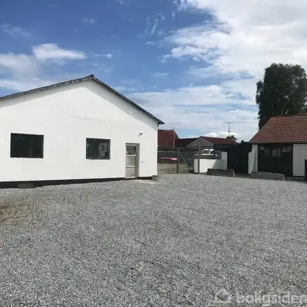 En hvid bygning står på en grusbelagt plads under en blå himmel med skyer. Omkring bygningen er der flere små huse og en stor træ i baggrunden.