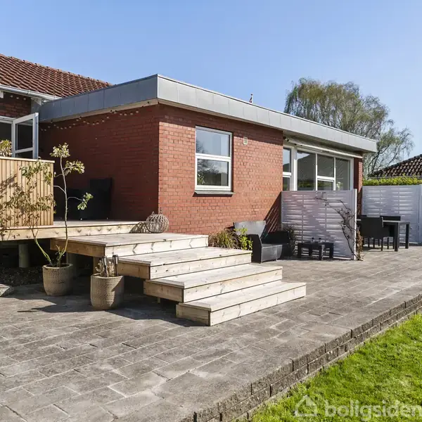 Rød murstensbygning med tilhørende træterrasse og flisebelagt gårdhave. Terrasse med planter i potter, og havemøbler i gårdhaven. Omringet af hække og andre huse i solrigt vejr.