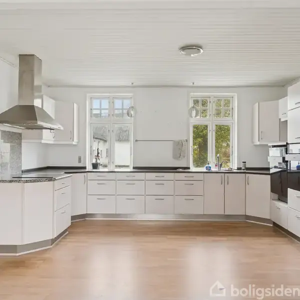 Lyst køkken med hvide skabe og sorte bordplader. Køkkenet har emhætte, komfur og indbyggede apparater. Rummelige vinduer slipper naturligt lys ind. Trægulv og hvidt loft fuldender rummet.