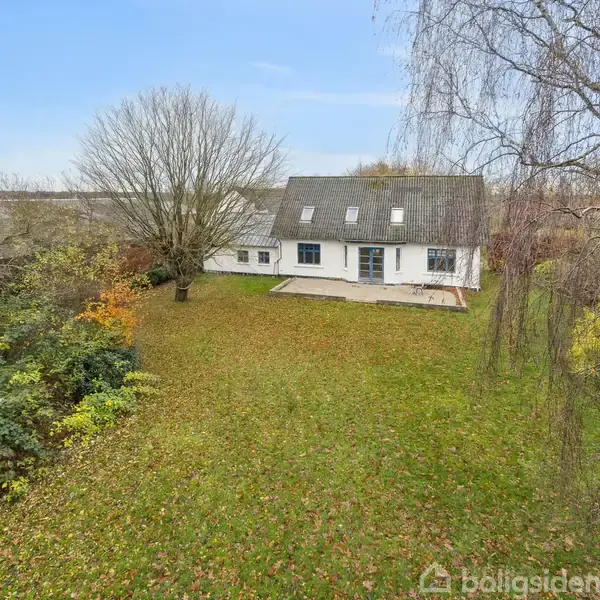 Et hvidt hus med skråt tag står midt i en stor, græsbeklædt have omgivet af træer og buske. Himlen er klar og blå.