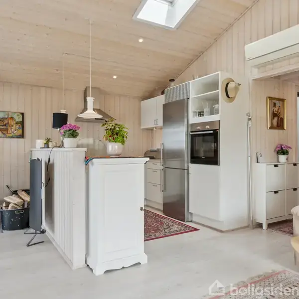 Komfur med brændestabel ved siden af står i hjørnet. Pejs ved siden af, brændende. Køkken med hvidt interiør, ovenlys i loftet, planter på bordet. Beige væg og gulv.