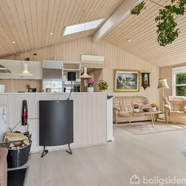 Et lyst, træbeklædt stueområde har en kamin til venstre, står i forgrunden. I baggrunden er en hyggekrog med lyse møbler og grønne planter, belyst af store vinduer.