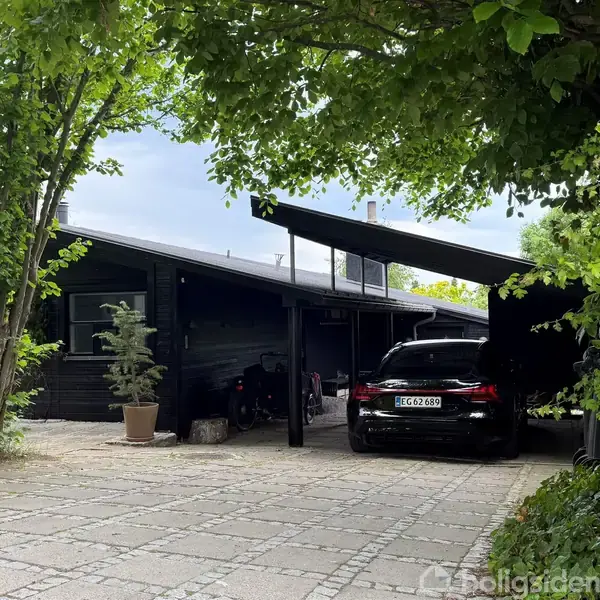 Sort træhus med store vinduer står på en hævet terrasse med moderne siddeområder. Træer og en klippet græsplæne omgiver terrassen.