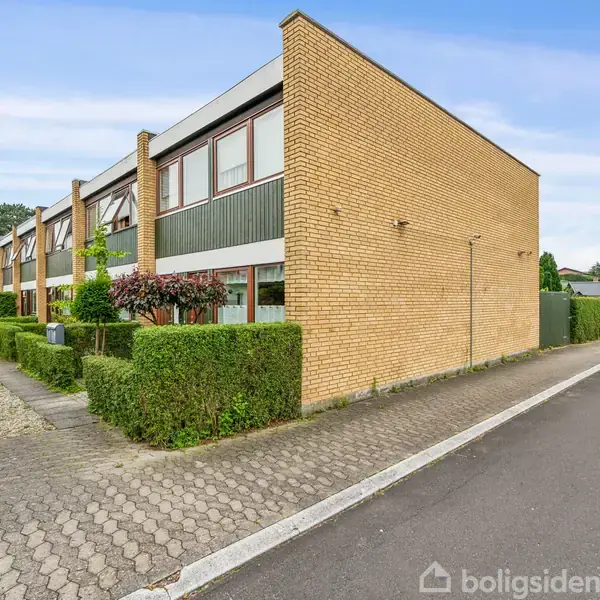 Rækkehus i gule mursten står langs en flisebelagt sti. Hækken omkring huset er veltrimmet. I baggrunden ses flere huse og grønne områder under en lys himmel.