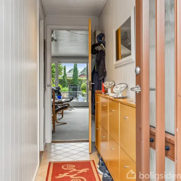 En smal entré har en orange skohylde med ure ovenpå. Til højre hænger frakker, en dør står åben, og i baggrunden ses en stue med store vinduer og træer udenfor.