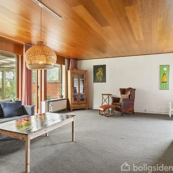 Stue med gråt tæppe har en blå hjørnesofa og et træsofabord. En stor flettet lampe hænger fra loftet. Baggrund indeholder en lænestol og vitrineskab.