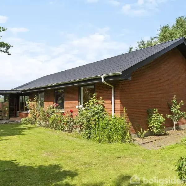 En rød murstensbungalow står omgivet af en grøn have, hvor buske vokser langs husmuren. I baggrunden ses træer og en klar blå himmel.