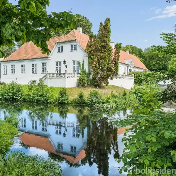 En hvid bygning med rødt tegltag står stille i en frodig, grøn have omkranset af træer og en lille å.